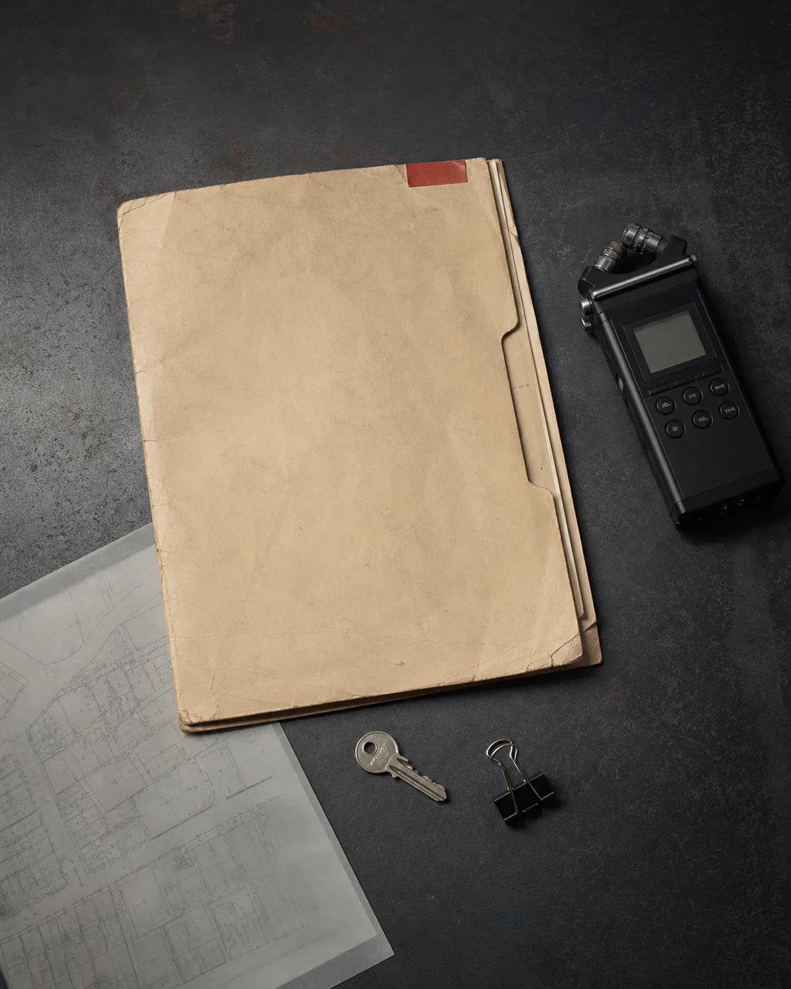 A manila case folder, field recorder, house key, and property map arranged on a charcoal desk.